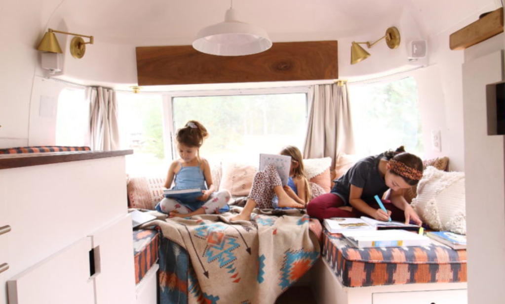 Three kids sit on a sunny bench seat in an airstream trailer with their schoolbooks open.